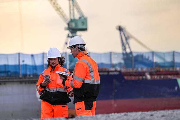 istock Two port engineers in high-visibility orange uniforms and hard hats communicate while inspecting dockside logistics using a laptop and walkie-talkie, symbolizing teamwork and safety. 2231741426