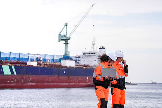 istock Two diverse engineers in safety gear coordinate a project at a waterside construction site. A female engineer reviews plans on a laptop while her colleague communicates on a radio. 2231742010