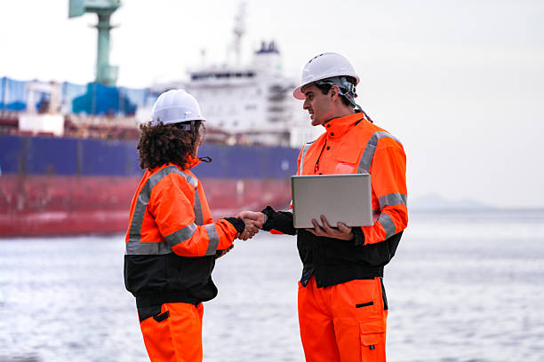 istock Two port engineers in high-visibility orange uniforms and hard hats communicate while inspecting dockside logistics using a laptop and walkie-talkie, symbolizing teamwork and safety. 2231742218