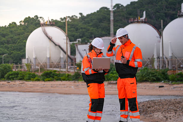 istock Two engineers in high-visibility safety gear and hard hats discuss project plans on a laptop in front of large spherical gas storage tanks at an industrial coastal facility. 2231742336