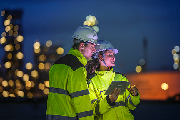 istock Two engineers in reflective safety gear review plans on a tablet during a night shift at an industrial site, with blurred factory lights glowing in the background. 2231742362