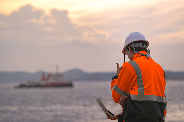 istock Two engineers wearing safety uniforms and hard hats communicate using a radio while reviewing data on a laptop by the sea during sunset. Maritime industrial setting. 2231744875