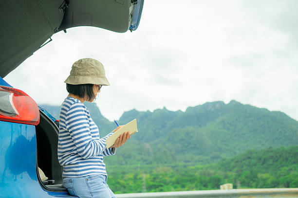istock Woman writing in notebook for digital detox. Self-reflection, journaling habit, and mindful planning beside blue car with mountain landscape background. Road trip travel lifestyle. Mindful travel. 2231753232