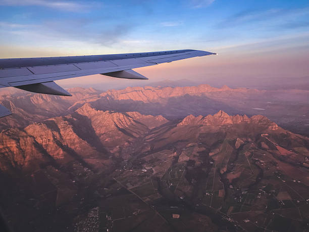 A breathtaking view of rugged mountain ranges during a flight, captured from a window seat at sunset. The airplane wing frames the scenery, complementing the warm hues of the golden hour sky. This image evokes a sense of exploration and the beauty of aerial perspectives while traveling.