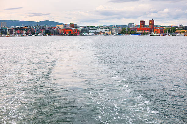 View of Oslo from the water with a visible boat wake leading toward the city skyline. The red brick Oslo City Hall and waterfront buildings stand against a backdrop of hills and cloudy sky