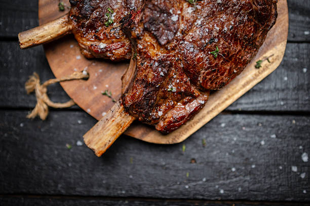 A close-up image of perfectly grilled tomahawk steaks served on a wooden cutting board, garnished with seasoning and herbs. The dish is presented against a dark textured background, emphasizing its rustic appeal. The juicy, crispy surface of the meat portrays a mouthwatering culinary masterpiece, perfect for promotions or presentations about fine dining and premium meals.