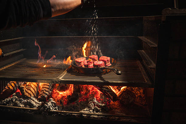 Juicy filet mignon cuts searing in a cast iron pan over an open flame grill. The glow of the fire and the seasoning being sprinkled create an appetizing ambiance perfect for culinary-focused content. This image captures the artistry of flame-cooked goodness.