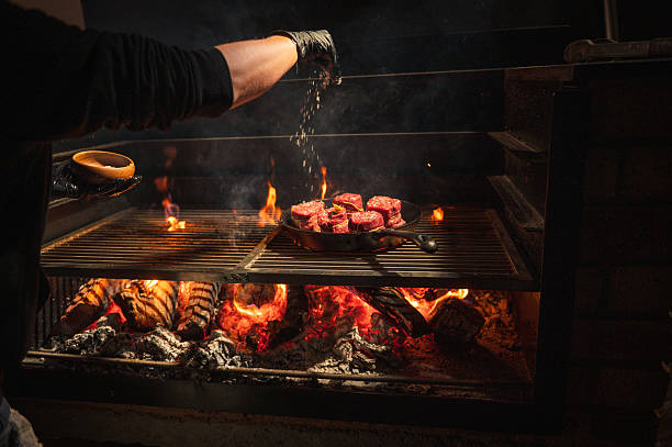 A skilled chef sprinkles seasoning onto perfectly cut steaks on a grill over open flames in a cozy ambiance. The image captures the art of cooking and grilling, symbolizing enjoyable meals and artisan culinary techniques. Complemented by the light of the fire, it conveys warmth and delectable flavors.