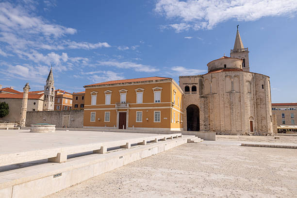 Ancient stone church with tall bell tower beside yellow building under blue sky. Historic Mediterranean town square with clear weather and open space. Architecture and urban landscape.