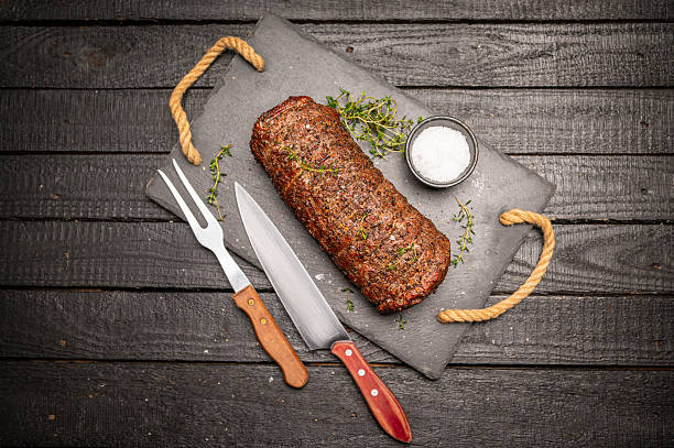 A roasted beef tenderloin garnished with fresh thyme is displayed on a slate serving tray. Accompanied by a seasoning bowl, a carving knife, and a fork, the setup rests on a dark, textured wooden surface, creating an atmosphere of rustic elegance and culinary craftsmanship.