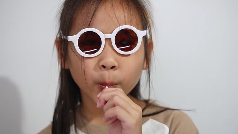 An Asian girl wearing round sunglasses while eating a lollipop, posing playfully with a fun and stylish expression against a white background.