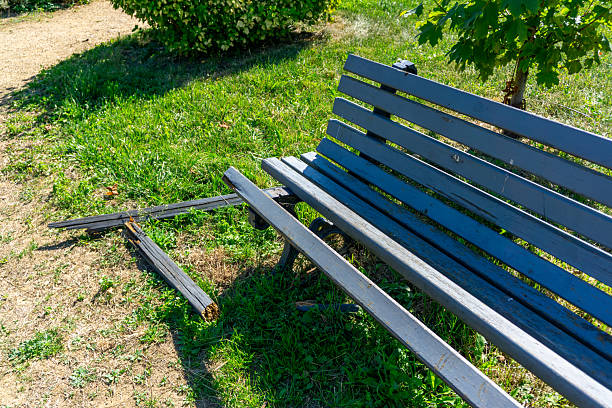 Broken park bench with detached wooden planks, damaged seat on grass surrounded by path and greenery.