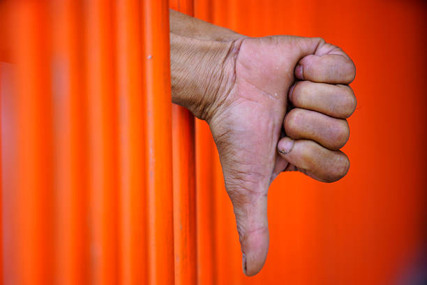 Hand with Thumbs Down Behind Orange Bars of Prison Cell Gate