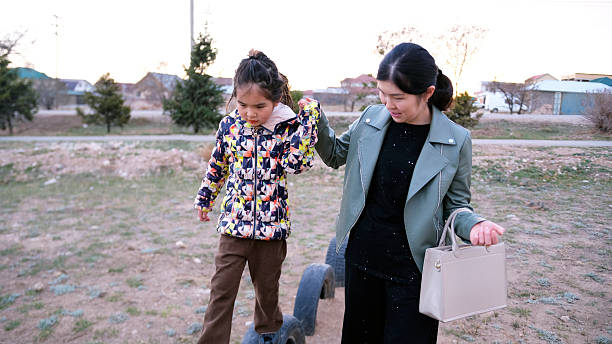 A single mom holds her little daughter's hand as she walks over tire tracks on a makeshift rural playground. A woman and a child are walking in the park