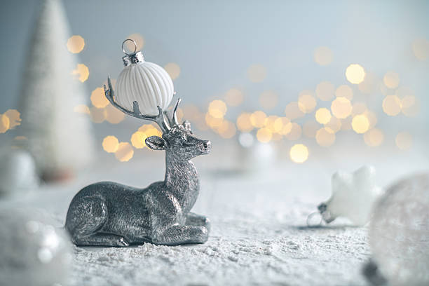 Silver reindeer figurine resting on snow-covered surface, surrounded by white christmas ornaments hanging amidst a backdrop of warm fairy lights, creating a magical and festive holiday atmosphere