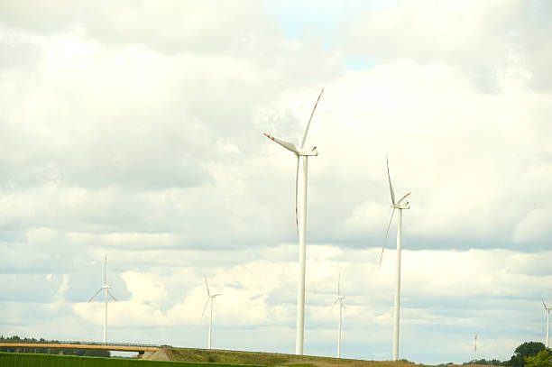 Wind turbines situated in an open field under a cloudy sky. The turbines are tall, white, and evenly spaced, capturing the wind efficiently with their elongated blades. A clear expanse of green land stretches beneath them. The sky is filled with large, white clouds, providing contrast to the turbines' sleek structures. A small section of a road or pathway is visible at the bottom left, indicating human infrastructure in the rural landscape.