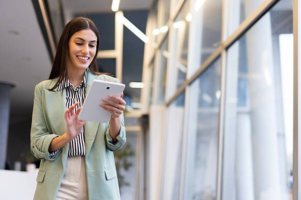 istock Young businesswoman smiling while walking through a modern office building, using a digital tablet and enjoying the bright, natural light 2231817180