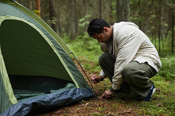 Caucasian young adult man setting up tent in forest, crouching on grass and securing tent peg, surrounded by trees and greenery, engaging in outdoor camping activity