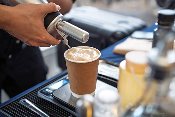 Close-up of a barista preparing a takeaway coffee in a paper cup, adding whipped cream topping using a dispenser.