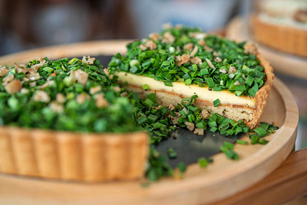 Close-up of a savory tart topped with fresh leeks, candied chicken ham and gruyere on a wooden serving tray, presented in a bakery setting.