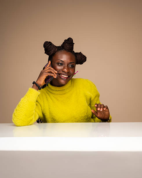 African woman in a bright yellow fuzzy sweater, styled with braided buns and pink floral earrings, smiles while holding a smartphone to her ear in a studio setting.