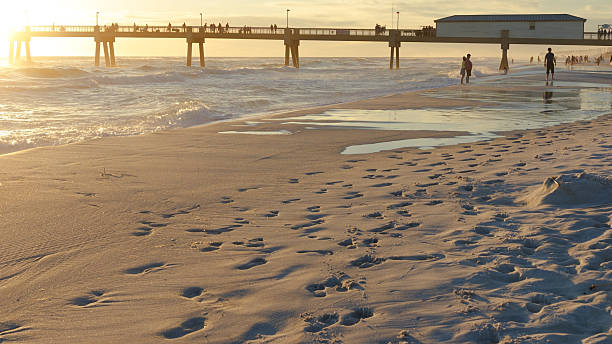Destin FL, United States - December 29, 2024: Beachgoers enjoy sunset strolls along sandy shore with pier silhouetted against golden sky