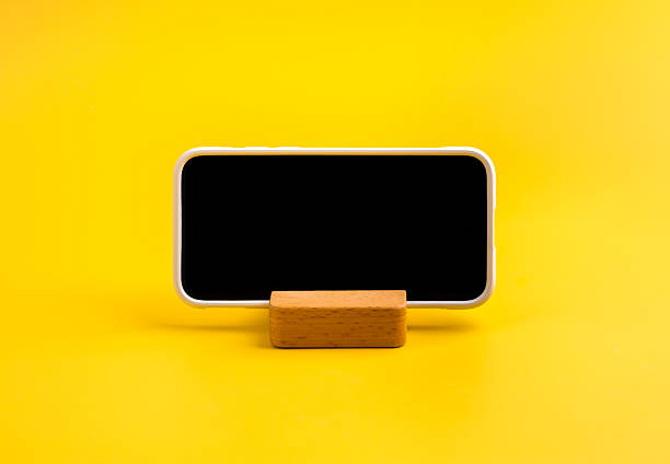 istock Smartphone with blank horizontal black screen on wooden stand on yellow background, concept of mobile technology, digital content, app design, mockup, online communication, UX and UI presentation. 2231909241