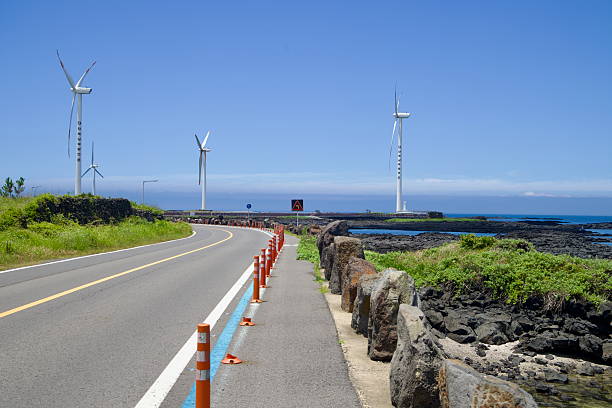 Orange lane bollards mark a curve on the Sinchang Windmill Coastal Road as tall turbines and basalt reefs line the bright blue shoreline.