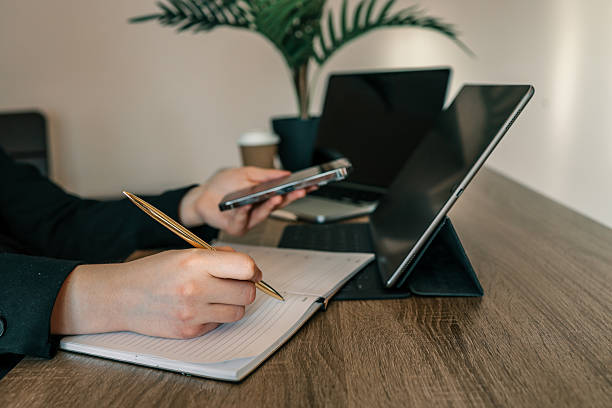 istock Person writing in notebook with gold pen while holding smartphone at desk with tablet, laptop, and coffee cup in modern workspace 2231918062