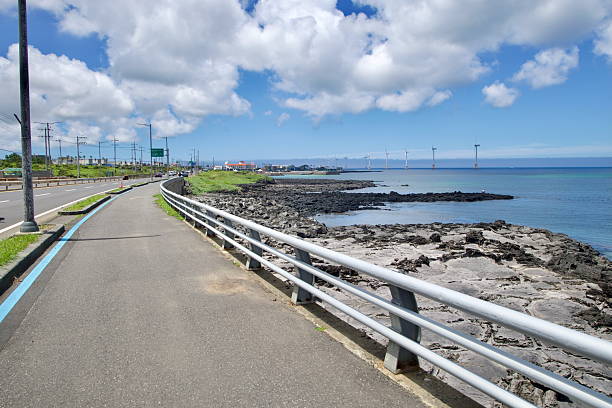 A bluelined bikeway parallels the coastal highway past basalt rocks and inlets toward Sinchang, with offshore wind turbines marking the skyline over calm water.