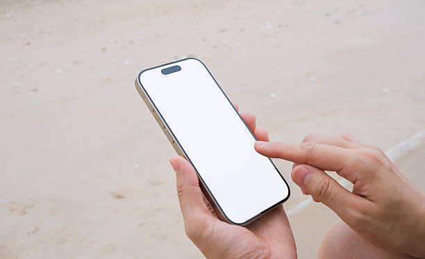 Hand holding a smartphone with a blank screen, on the sea and sandy beach background. Summer vacation concept.