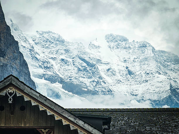 Snow-covered peaks of the Swiss Alps rise dramatically behind traditional chalet rooftops in Lauterbrunnen, Switzerland. The photo captures the contrast between the rustic wooden architecture in the foreground and the towering alpine mountains in the background, partially veiled by clouds.