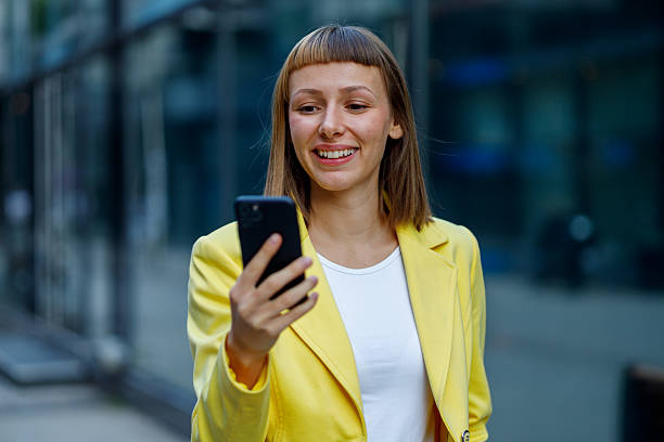 istock Young businesswoman wearing a vibrant yellow jacket, smiling while using her smartphone in a bustling urban cityscape 2231939295
