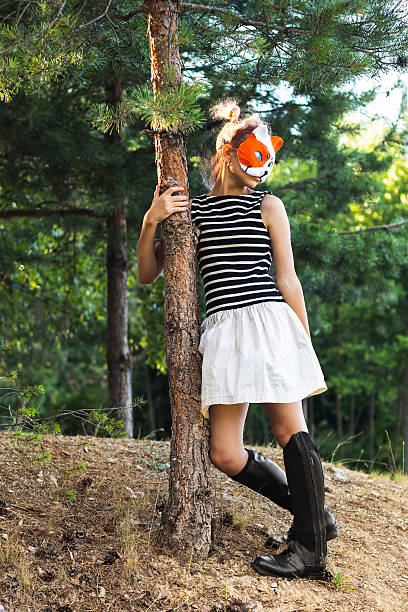 A young girl, part of the quadrobics subculture, poses playfully in a fox mask and boots against a tree in a sunny forest setting. She embodies the spirit of animal movement and connection to nature.