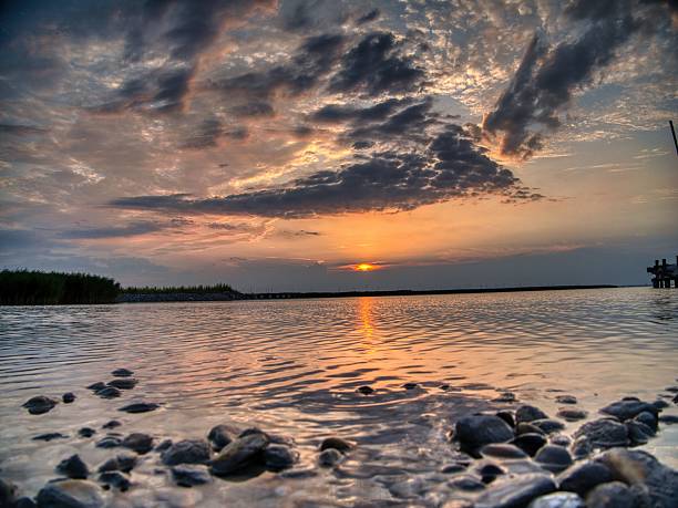 A serene sunset over a calm lake with rocky shore and dramatic clouds in the sky.