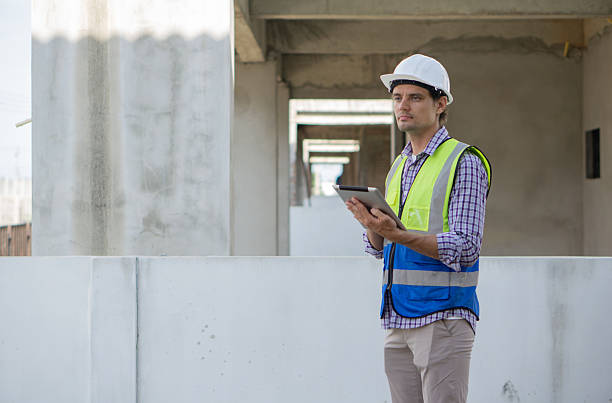 istock Young caucasian worker in hardhat and safety vest review plan on tablet computer. Working atmosphere at a construction site with unfinished concrete wall. 2231954832