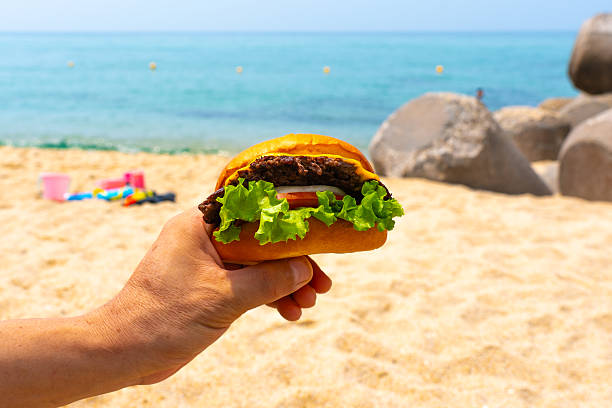 istock Man holding burger in a hand against beach background 2231966441