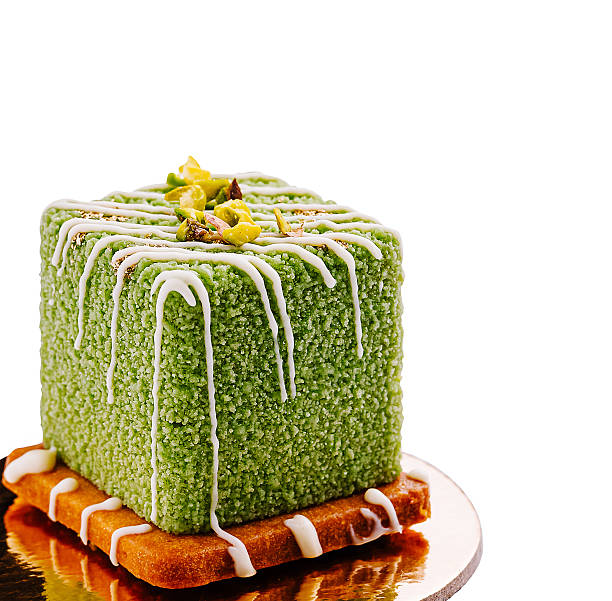 This dessert features a vibrant green cube layered on a golden crust. It is topped with a drizzle and sprinkled with nuts, showcasing an artistic culinary creation.