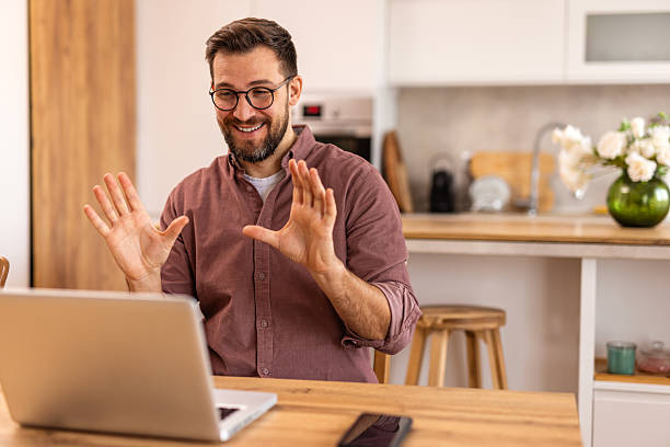 istock Friendly man greeting someone on a video call while working from home. He sits in a stylish kitchen with a laptop, orange juice, and warm lighting, creating a welcoming and professional setting. 2231969627
