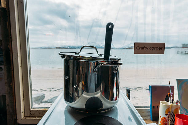 Large stainless steel pot with lid and spoon on stove by window overlooking sandy beach and ocean. Kitchen scene with cooking utensils and craft supplies nearby. Coastal home interior.