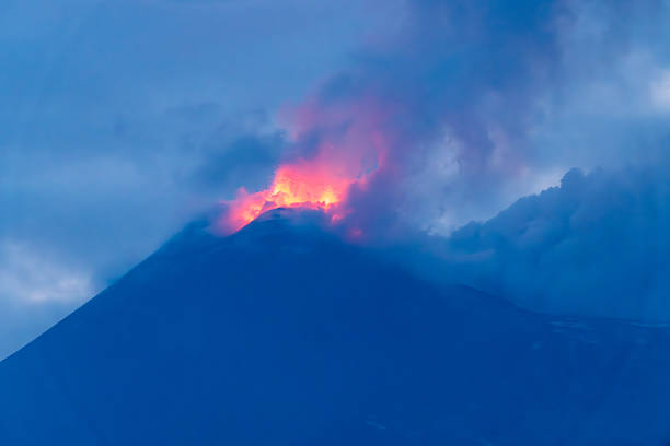 Volcanic eruption emits glowing lava and dense smoke into cloudy sky during daytime, partially obscuring the summit of Mount Etna.
