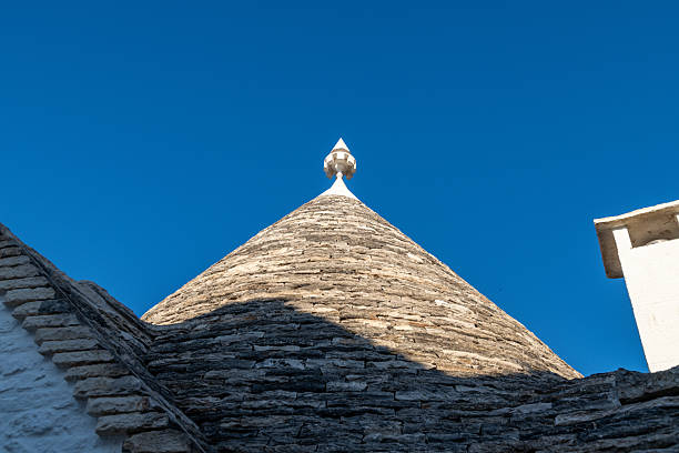 A detailed view of a traditional conical stone roof with white finial against a deep blue sky, captured in the town of Alberobello in Italy.
