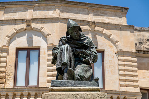 A seated bronze statue in armor is shown in front of a stone building. Location: Lecce - Italy Piazzetta Raimondello Orsini Lecce.