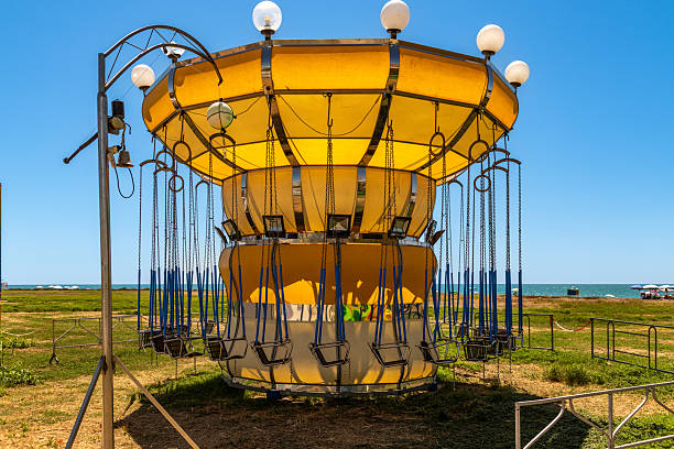 A yellow swing carousel stands unused on a grassy area near the sea, surrounded by fences, lighting, and a vibrant blue summer sky.