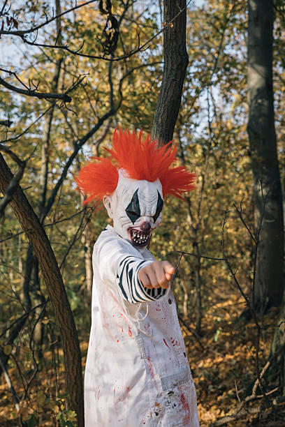 Child wearing a scary clown mask and bloody costume pointing finger in autumn forest during Halloween