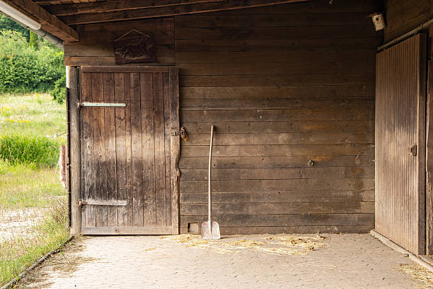 A weathered horse stable at the edge of a meadow, capturing the essence of rural outdoor life. The aged wooden walls bear the marks of time and exposure, while a half-open stable door leads out to the lush countryside. A well-used pitchfork leans against the wall amid scattered straw, evoking the quiet rhythm of daily farm work. This image reflects the simplicity and raw beauty of life on the land.