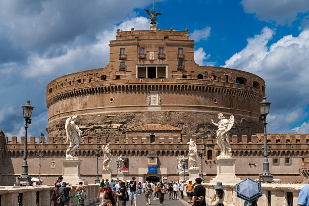 Rome - Lazio - Italy - 08-02-2025: Tourists walk across the bridge lined with statues leading towards the historic Castel SantAngelo under a cloudy blue summer sky.