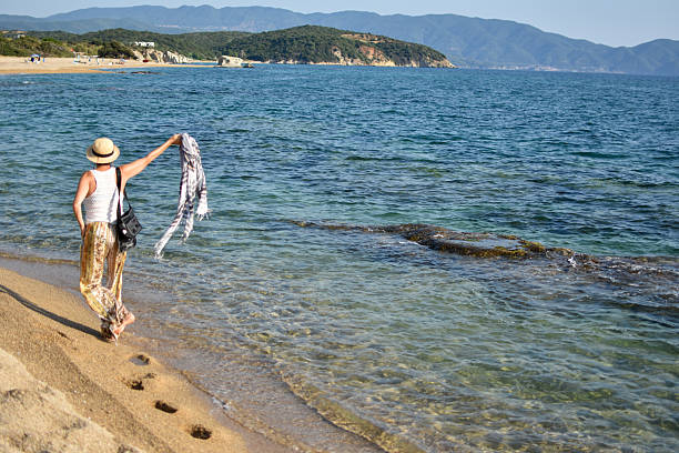 middle-aged woman walking on a sandy beach on a nice sunny day, casually dressed, wearing a hat, holding a scarf in her outstretched hand, blowing in the wind, the sea is visible, her back is turned