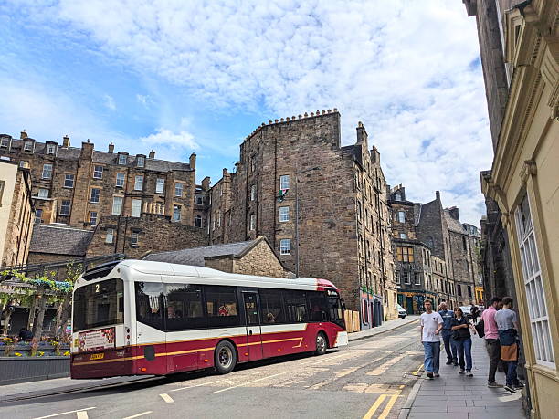 Edinburgh, Scotland - July 17 2025: A modern public bus travels along a narrow historic Candlemaker Row as pedestrians walk on the sidewalk in the Old Town under a partly cloudy sky on a summer day