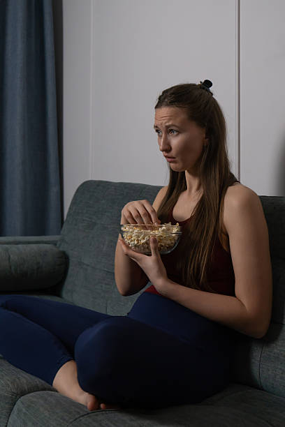 istock Young woman sitting on a sofa at home, enjoying popcorn from a glass bowl while watching a movie or tv show with a serious and focused expression, immersed in entertainment. Vertical photo 2231990756